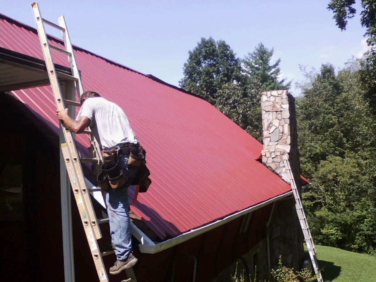 Roofer on a ladder beside a red metal roof and stone chimney — Soffit & Fascia Repair by Roofing Brothers in Poolville